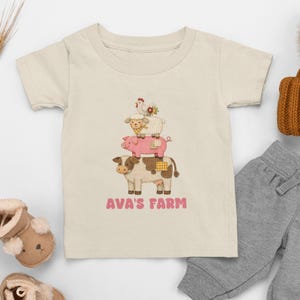 Peut inclure: T-shirt crème pour enfant avec un motif d'animaux de la ferme empilés. Une vache, un cochon, un mouton et une poule sont superposés. Le texte "Ava's Farm" est imprimé en rose. Le t-shirt est associé à un pantalon de survêtement gris et un bonnet marron.