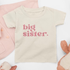 May include: A cream coloured t-shirt with short sleeves and the words "big sister" printed in pink.