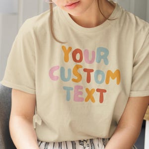 May include: A cream-coloured t-shirt featuring the text "YOUR CUSTOM TEXT" in colourful bubble letters. The text is in orange, pink, blue, and red. The shirt has short sleeves.
