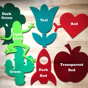 May include: Assorted laser-cut shapes in various colors, including a dark green tree, a light green frog, a green cactus, a teal flower, a red heart with an arrow, a dark red rocket, and a transparent red apple. Each shape has a color label.