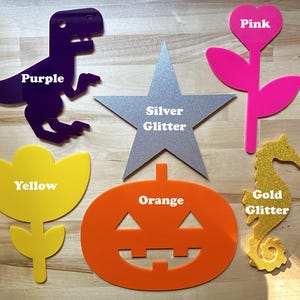 May include: Assortment of colorful acrylic shapes, including a purple dinosaur, a silver glitter star, a pink flower, a yellow flower, an orange pumpkin, and a gold glitter seahorse. Each shape has a label with its color.