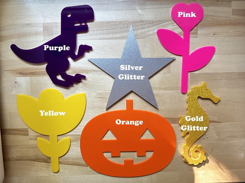 May include: Assortment of colorful acrylic shapes, including a purple dinosaur, a silver glitter star, a pink flower, a yellow flower, an orange pumpkin, and a gold glitter seahorse. Each shape has a label with its color.