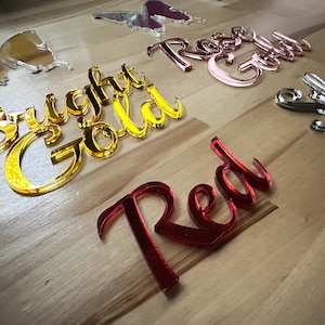 May include: A collection of decorative acrylic words and butterfly shapes in various colors. The words include "Bright Gold", "Rose Gold", "Red", and "Silver". The butterfly shapes are gold and clear.