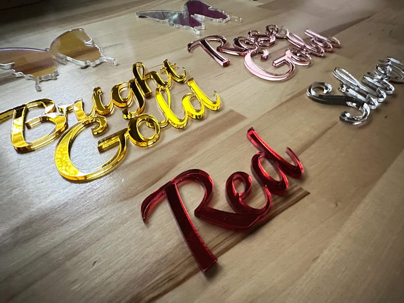 May include: A collection of decorative acrylic words and butterfly shapes in various colors. The words include "Bright Gold", "Rose Gold", "Red", and "Silver". The butterfly shapes are gold and clear.