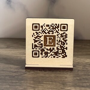 Peut inclure: Un support en bois avec un code QR gravé au laser et la lettre "E" au centre. Le support est en bois brun clair et a une base brun foncé.