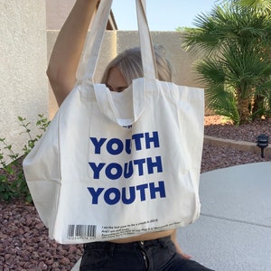 Pu&ograve; includere: Una borsa di tela bianca con testo blu che dice "YOUTH" tre volte. La borsa ha una breve descrizione di testo che dice "I am the last year to be a youth in 2019. And I am still here. Someday, I will be able to say that it is "Not youth not older." We were born to grow."