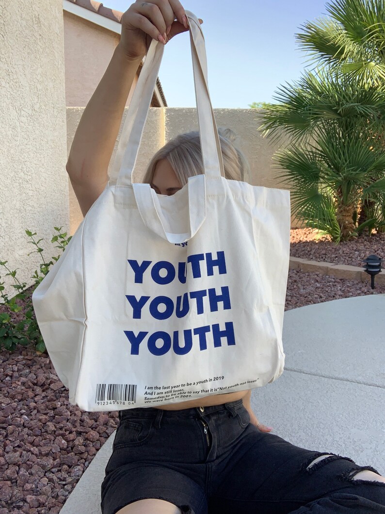 Pu&ograve; includere: Una borsa di tela bianca con testo blu che dice "YOUTH" tre volte. La borsa ha una breve descrizione di testo che dice "I am the last year to be a youth in 2019. And I am still here. Someday, I will be able to say that it is "Not youth not older." We were born to grow."