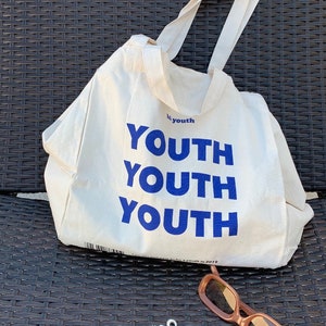 Pu&ograve; includere: Una borsa tote in tela bianca con testo blu che recita "by youth YOUTH YOUTH YOUTH". La borsa &egrave; appoggiata su una superficie in vimini intrecciato.