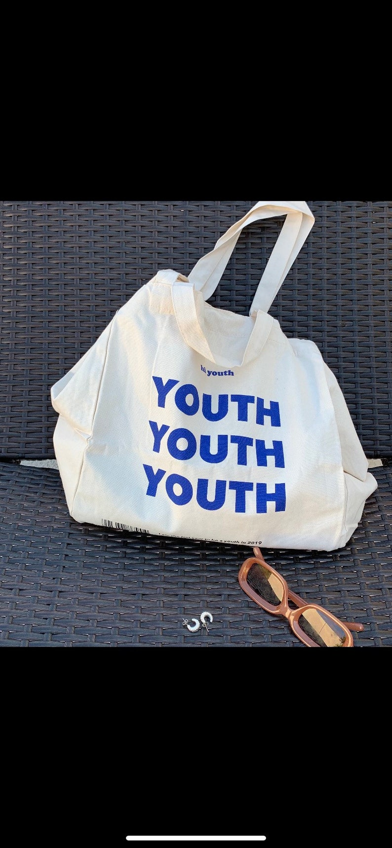 Pu&ograve; includere: Una borsa tote in tela bianca con testo blu che recita "by youth YOUTH YOUTH YOUTH". La borsa &egrave; appoggiata su una superficie in vimini intrecciato.