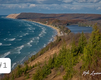 May include: An aerial view of a sandy beach with blue water and waves. The beach is lined with trees and a small lake is visible in the distance. The text "#1" is in the lower left corner of the image.
