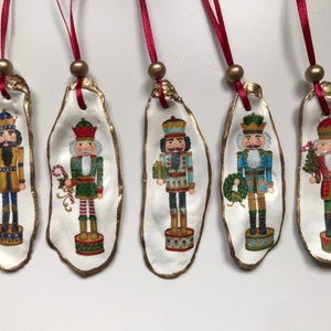 May include: Five Christmas tree ornaments made from oyster shells, each decorated with a painted nutcracker soldier. The shells have gold edging, red ribbon hangers, and gold beads. The nutcrackers are in different colours.