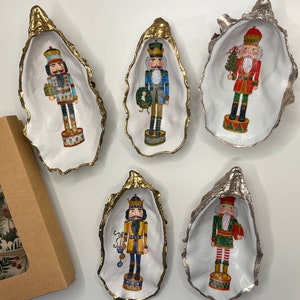 May include: Five oyster shell trinket dishes with gold and silver metallic edges, each featuring a colourful nutcracker soldier design. A gift box with a holiday pattern is visible on the left.