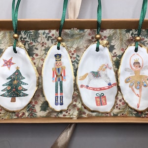 May include: A set of four Christmas oyster shell ornaments. Each shell features a festive design: a Christmas tree, a nutcracker, a rocking horse, and a ballerina. Gold edging and green ribbon for hanging.