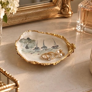 May include: An oyster shell trinket dish with a gold-painted rim and a hand-painted watercolour illustration of three sailboats on the water. The dish contains two gold rings and a pair of small white pearl stud earrings.