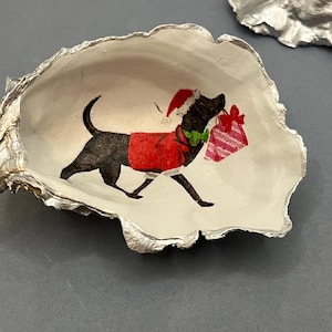 May include: An oyster shell with a silver trim, featuring a black dog illustration in a Santa hat and red coat, carrying a gift. The shell is on a grey surface. A festive decorative item.