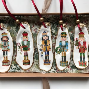 May include: Five Christmas tree ornaments with nutcracker illustrations on oyster shells. Each shell has a gold edge and a red ribbon. The ornaments are presented in a decorative box, ideal for festive decor.
