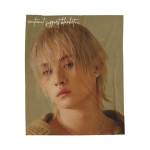 May include: A blanket with the text "emotional support blanket..." is displayed. The blanket features a person with blonde hair and a neutral expression. The background is a muted, warm tone.