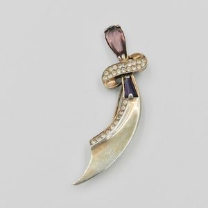 May include: A silver and gold tone brooch with a crescent shape and a purple and clear rhinestone design.