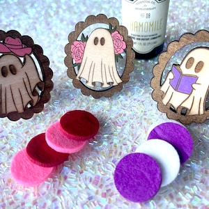 May include: Three wooden ghost ornaments with different designs. The first ghost is wearing a pink cowboy hat, the second ghost has pink flowers around it, and the third ghost is reading a purple book. The ornaments are on a white sparkly surface with pink and purple felt circles.
