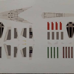 Könnte beinhalten: Ein Satz weißer und grauer Abziehbilder für ein Raumschiffmodell. Die Abziehbilder beinhalten ein langes, spitzes Schiff, ein Cockpit und verschiedene andere Details. Die Abziehbilder sind in zwei Reihen angeordnet, mit dem Schiff oben und den anderen Details darunter. Die Abziehbilder sind auf weißem Hintergrund gedruckt.