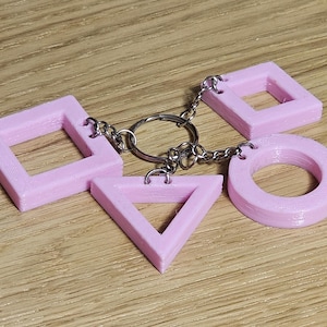 Può includere: Un portachiavi rosa con quattro forme geometriche: un quadrato, un triangolo, un cerchio e un quadrato. Le forme sono collegate da catene argentate e un portachiavi. Il portachiavi è realizzato in un materiale rosa chiaro.