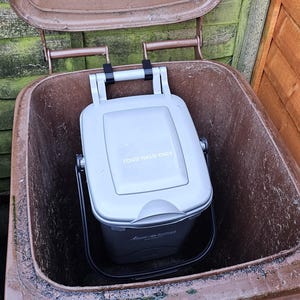Op de afbeelding: Een grijze compostbak met een handvat staat in een bruine buitencompostbak. De bak heeft een scharnierend deksel en de woorden "Food Waste Bin" zijn zichtbaar. Links is gedeeltelijk een blauwe recyclingbak te zien.
