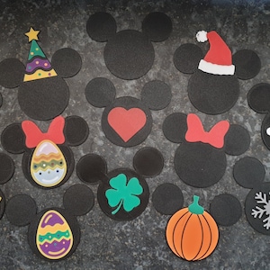 Könnte beinhalten: Ein Set mit 14 schwarzen Filz-Mickey-Mouse-Ohren mit verschiedenen Feiertags- und Saison-Designs. Die Designs umfassen einen Weihnachtshut, einen Regenbogen, ein Kleeblatt, einen Kürbis, eine Schneeflocke, ein Herz, eine Narzisse, ein Osterei, einen Hasen, einen Partyhut und eine Schleife.