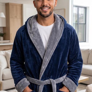 May include: A plush navy blue bathrobe featuring a contrasting light gray interior lining on the hood, cuffs, and front trim. The robe includes a matching tie belt at the waist and two large patch pockets on the front, worn over a white t-shirt.