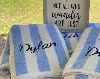 May include: Two white and blue striped beach towels with the names "Dylan" and "Drake" embroidered on them. The towels are folded and stacked on a wooden table. A sign above the towels reads "Not all who wander are lost".