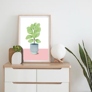 May include: A framed print of a green leafy plant in a blue and white patterned pot on a pink background. The print is on a wooden shelf with a small potted plant and a white lamp.