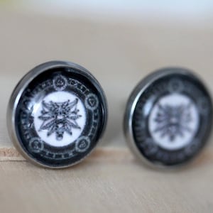 Può includere: Un paio di orecchini a bottone d'argento con un design in bianco e nero. Il design presenta una testa di lupo circondata da un motivo circolare.