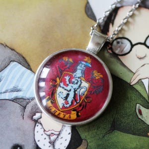 Peut inclure: Un collier avec un pendentif rond en argent orné d'un blason rouge et or avec un griffon bleu. Le pendentif porte l'inscription "GRETA" en lettres dorées. L'arrière-plan comprend une illustration de dessin animé.