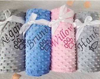 Pode incluir: Quatro mantas de pontos minky enroladas em cinza, azul, rosa e cinza claro. Cada manta é amarrada com uma fita de cetim branca e apresenta nomes bordados: Reggie, Bruno, Willow e Hank, acompanhados por ícones de patas ou cenouras.