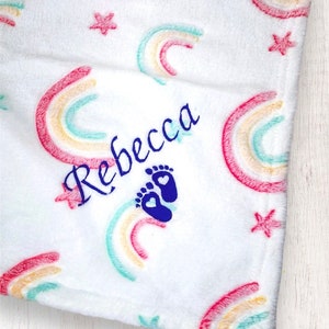 May include: White baby blanket with a rainbow pattern and stars. The blanket is personalized with the name "Rebecca" and a blue footprint with a heart.