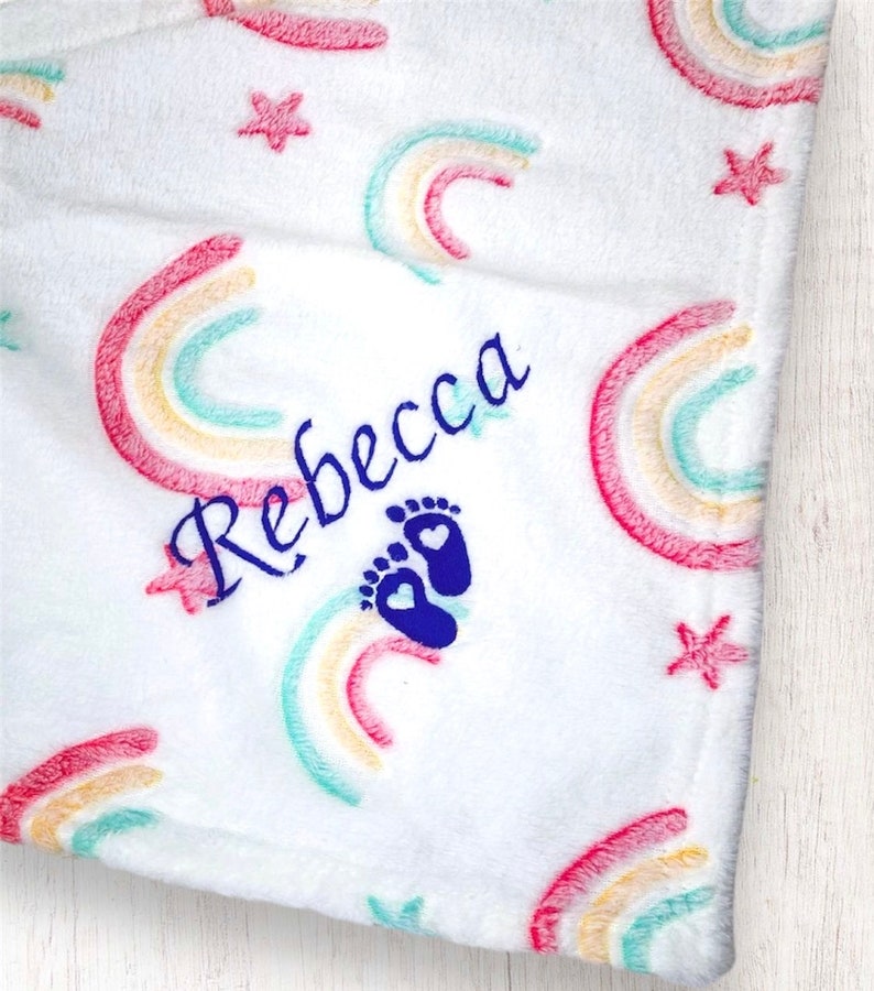 May include: White baby blanket with a rainbow pattern and stars. The blanket is personalized with the name "Rebecca" and a blue footprint with a heart.