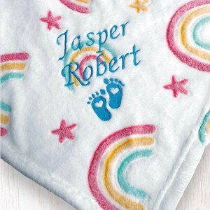 May include: White baby blanket with embroidered rainbows, stars, and footprints. The blanket is personalized with the names "Jasper Robert".