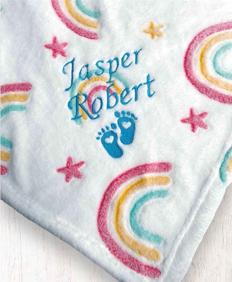 May include: White baby blanket with embroidered rainbows, stars, and footprints. The blanket is personalized with the names "Jasper Robert".
