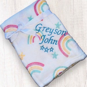 May include: White baby blanket with a rainbow and star pattern. The blanket is tied with a white ribbon and has the name "Greyson John" embroidered on it.