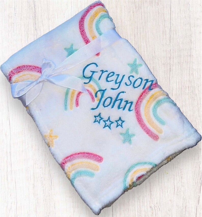 May include: White baby blanket with a rainbow and star pattern. The blanket is tied with a white ribbon and has the name "Greyson John" embroidered on it.