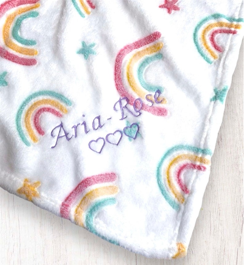 May include: White plush blanket with a rainbow pattern and stars. The blanket is personalized with the name "Aria-Rose" and three hearts.