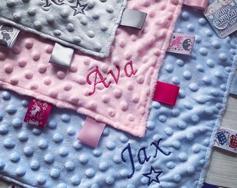 Può includere: Tre copertine di sicurezza quadrate per neonati in grigio, rosa e azzurro con una texture minky a pois. Ogni copertina presenta i nomi ricamati Ronnie, Ava e Jax con motivi a stella, insieme a vari nastri colorati con motivi decorativi.