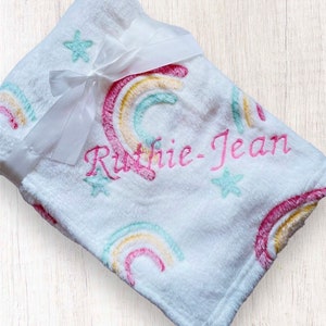 May include: White plush blanket with a rainbow and star pattern. The blanket is personalized with the name "Ruthie-Jean" embroidered in pink.