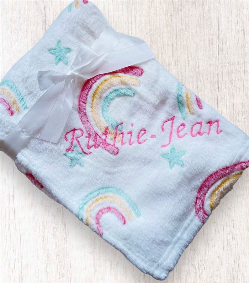 May include: White plush blanket with a rainbow and star pattern. The blanket is personalized with the name "Ruthie-Jean" embroidered in pink.