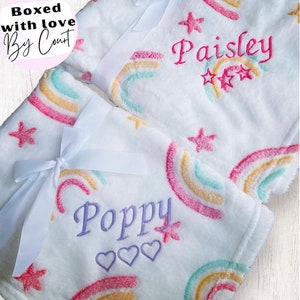 May include: Two white plush blankets with rainbow and star designs. The blankets are personalized with the names "Poppy" and "Paisley".
