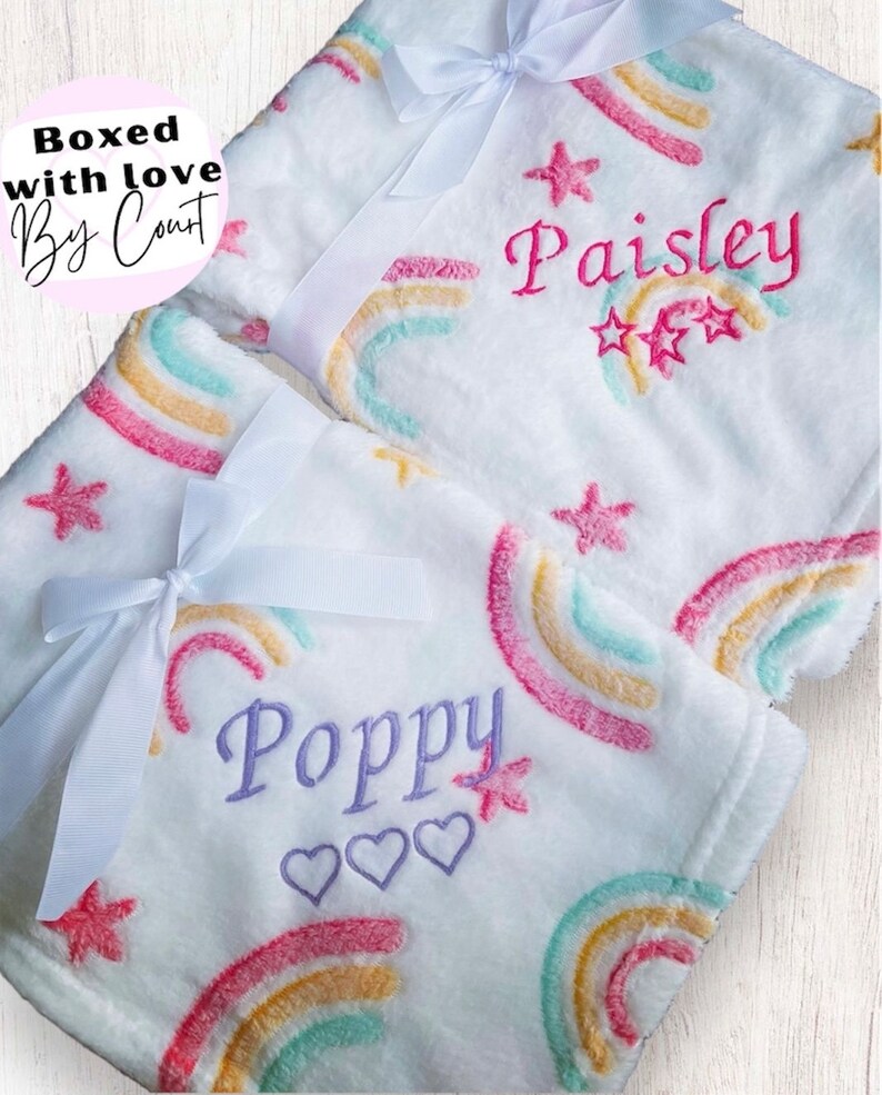 May include: Two white plush blankets with rainbow and star designs. The blankets are personalized with the names "Poppy" and "Paisley".