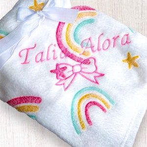 May include: White plush baby blanket with a pink bow, rainbow design, and the name "Tali Alora" embroidered in pink.