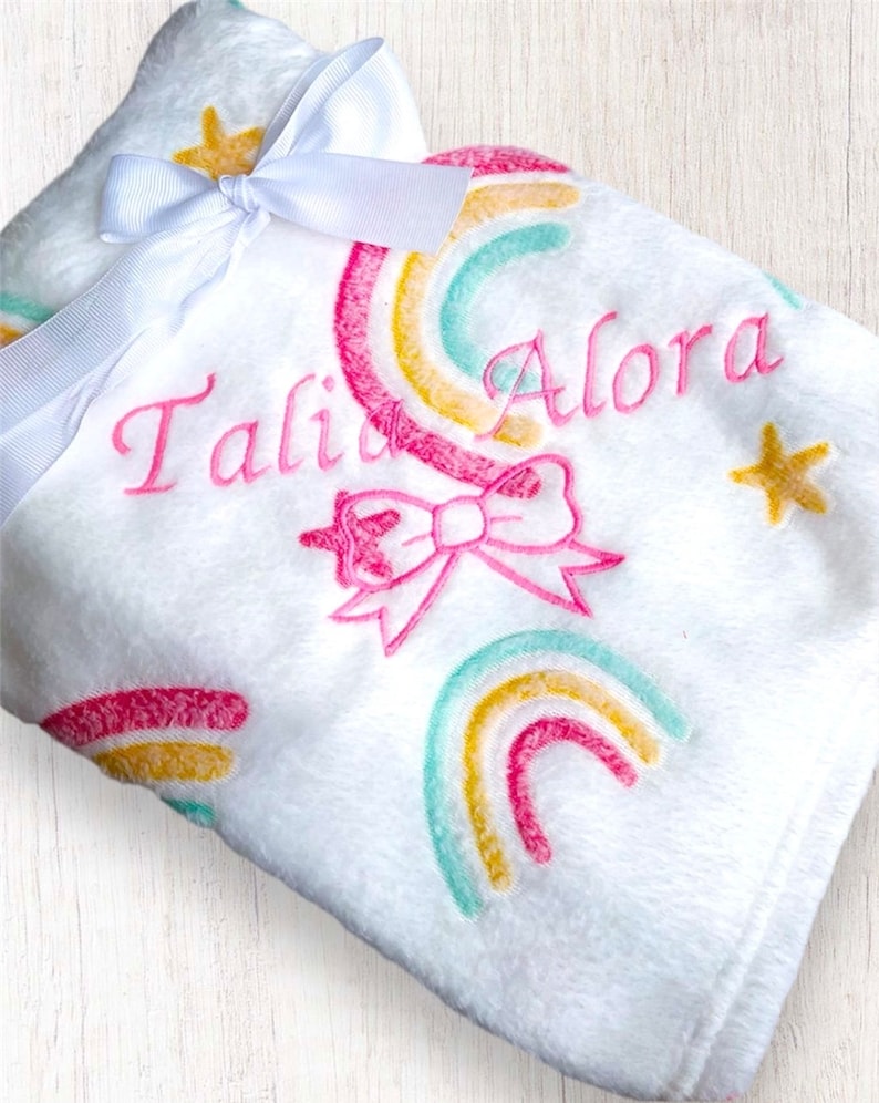 May include: White plush baby blanket with a pink bow, rainbow design, and the name "Tali Alora" embroidered in pink.