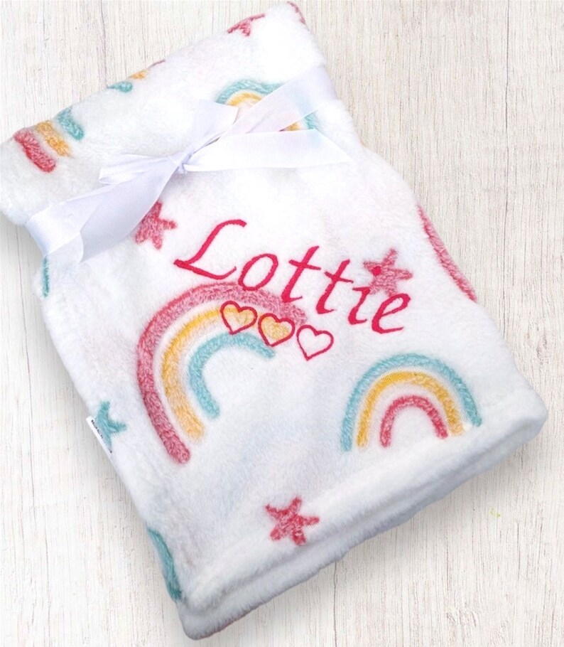May include: White fleece baby blanket with a rainbow and star pattern. The blanket is personalized with the name "Lottie" in pink embroidery.