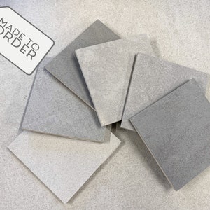 May include: Six square tiles in various shades of grey, from light to dark, arranged in a pile on a white surface. The tiles have a textured surface and a slightly rough finish. A tag with the text "MADE TO ORDER" is visible in the top left corner of the image.