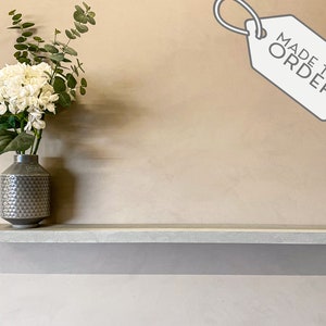 May include: A grey concrete floating shelf on a beige wall. A grey vase with a geometric pattern holds white hydrangeas and eucalyptus. A tag with "MADE TO ORDER" is in the upper right.